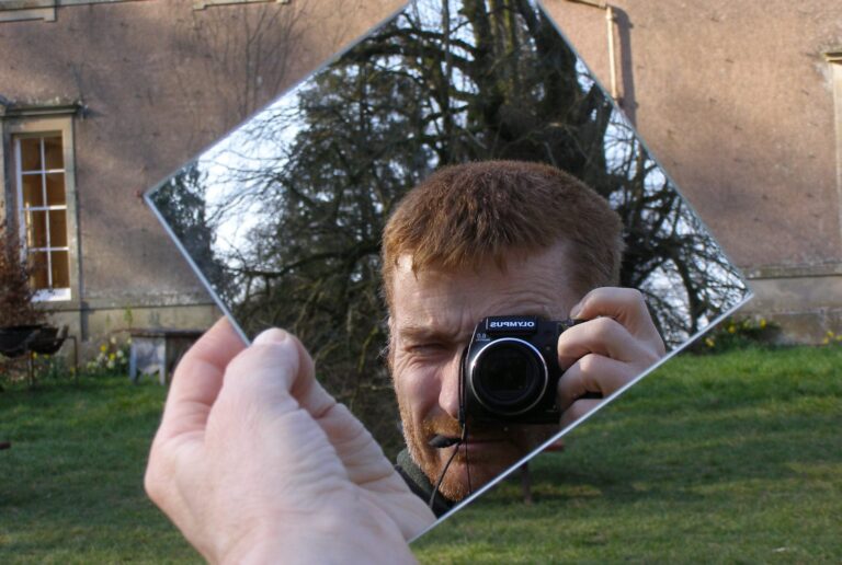 Photographer and trees reflected in a square mirror, with a large house behind